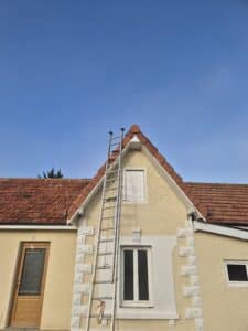 Façade de maison jaune, toit en tuiles rouges sous ciel bleu. Échelle en aluminium dressée sur le pignon pour travaux. Pistolet à calfeutrer suspendu.